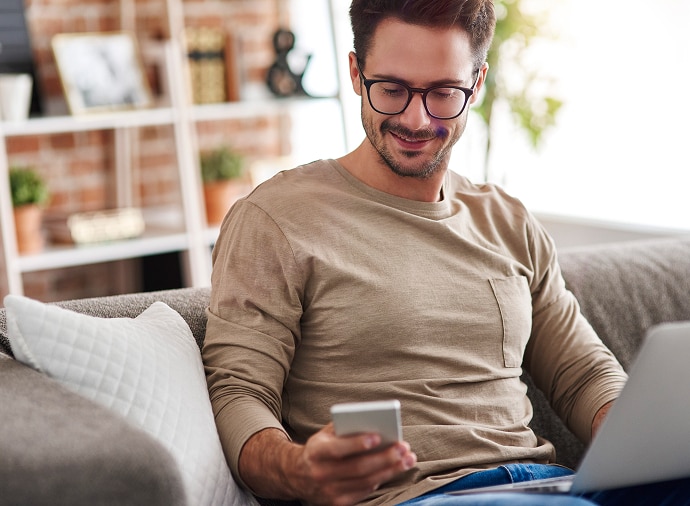 Man zit op de bank en kijkt op zijn telefoon terwijl hij een laptop op schoot heeft